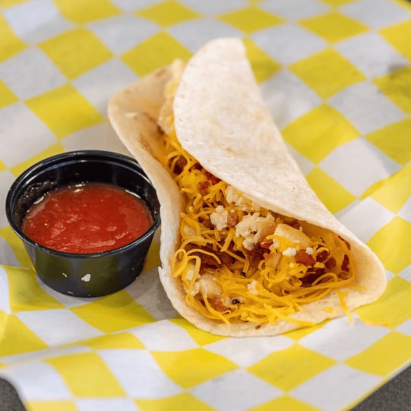 A Casa del Taco breakfast taco with cheese, bacon, and eggs, served with salsa on checkered paper.