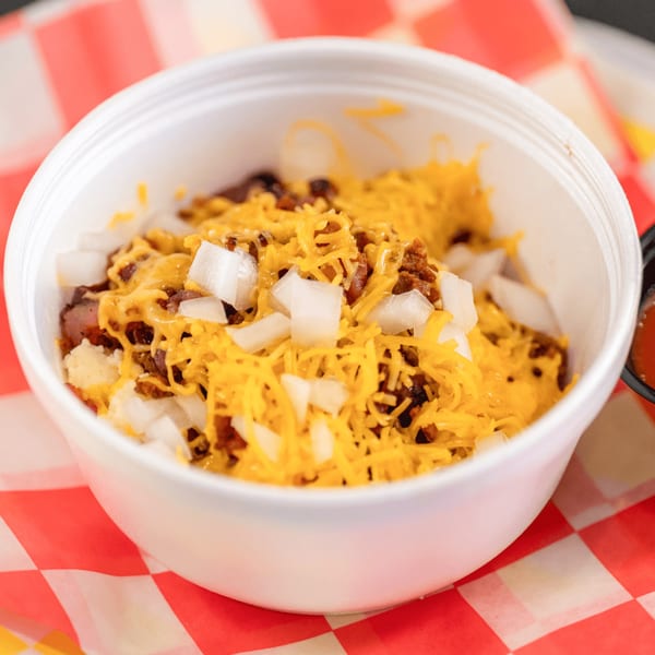 A white bowl of Casa del Taco chili with shredded cheese and diced onions on a red and white checkered paper.