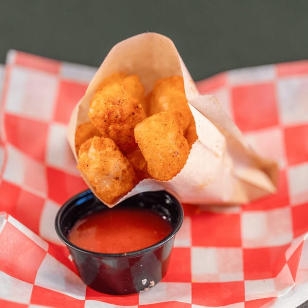 Crispy golden tater tots in a paper bag with a side of red dipping sauce on a checkered liner.