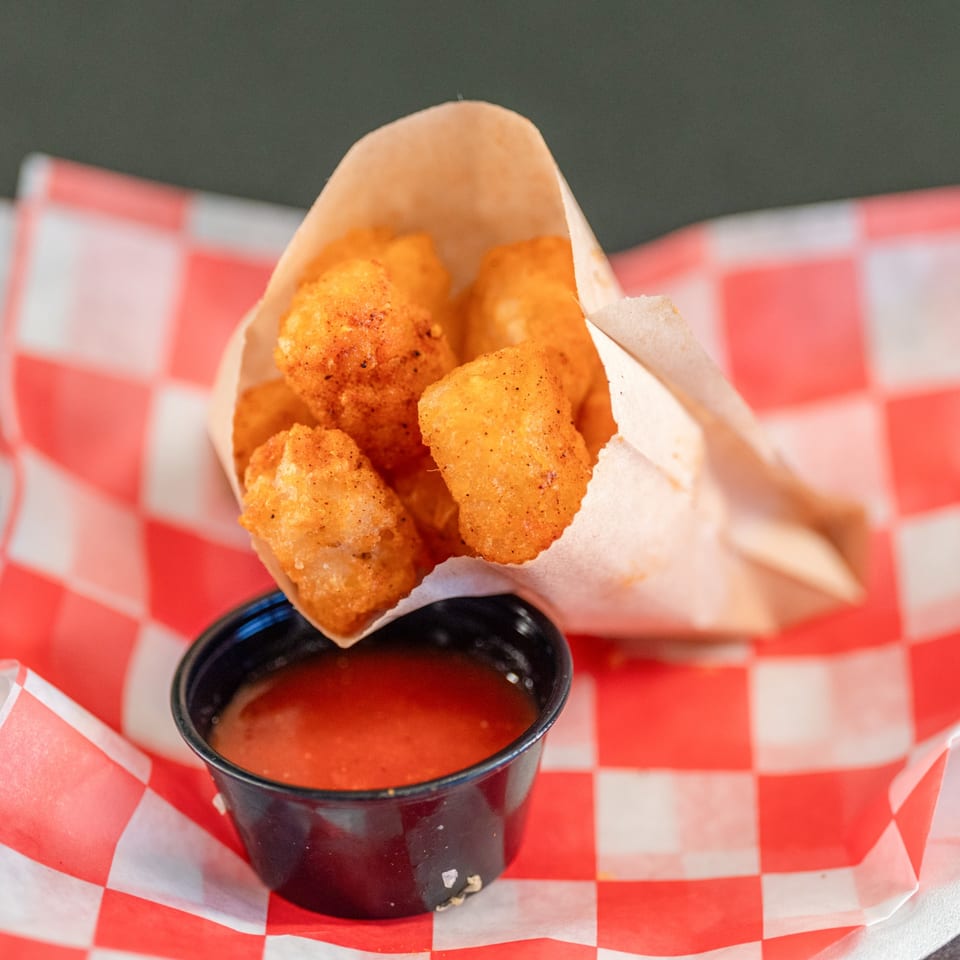 Golden tater tots in a paper cone with a side of red dipping sauce on a red and white checkered liner.