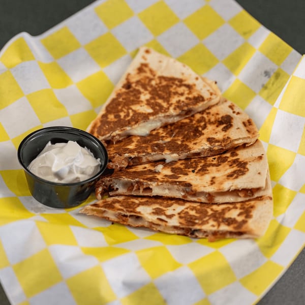 Four quesadilla slices with melted cheese and a side of sour cream on yellow checkered paper.