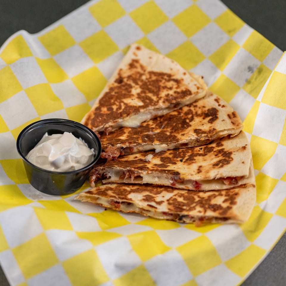 Four quesadilla slices with melted cheese and meat, served with a side of sour cream on checkered paper.